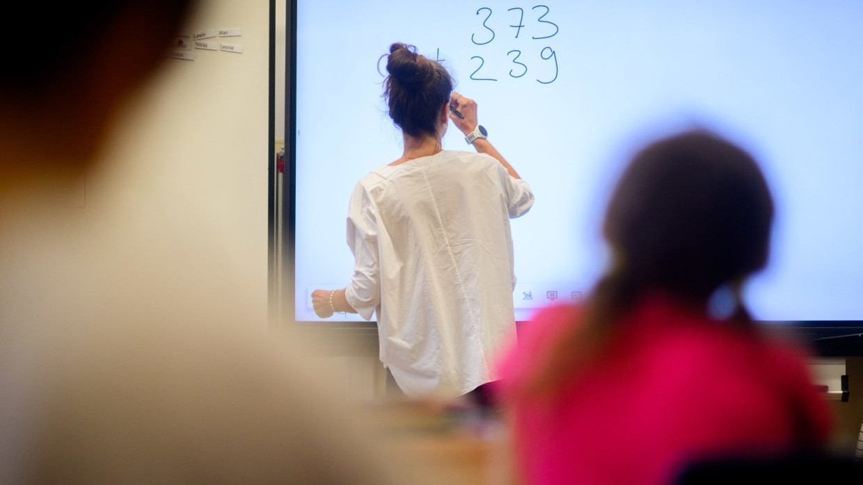 Eine Lehrerin schreibt eine Mathematikaufgabe auf eine digitale Schultafel im Klassenraum einer 4. Klasse einer Grundschule.